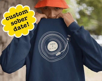 May include: Navy blue sweatshirt featuring a white graphic of a record player with the word "SOBER" and the date "01.12.20". A yellow flower-shaped sticker with the text "custom sober date!" is in the upper left corner.
