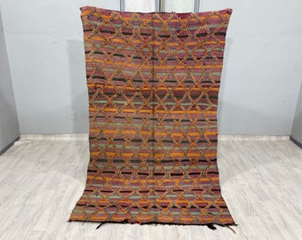 Puede incluir: Una alfombra vintage con un patrón geométrico en tonos naranja, morado, verde y negro. La alfombra tiene un patrón de diamante con un borde de flecos.