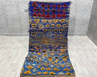 May include: A rectangular, handmade rug with a vibrant blue background and geometric patterns in orange, black, and red. The rug features a textured weave and a fringed edge, adding to its rustic charm. The design includes diamond and floral motifs.