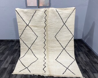 May include: A white Moroccan rug with a black diamond pattern. The rug has a fringe border and is made of wool.