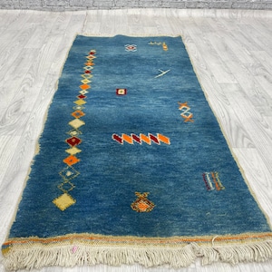 Puede incluir: Una alfombra azul con un patrón geométrico en tonos naranja, rojo y amarillo. La alfombra tiene un borde de flecos.