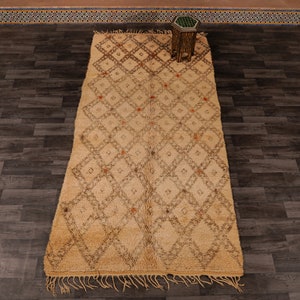 Pu&ograve; includere: Un tappeto marocchino beige con un motivo a rombi marrone. Il tappeto ha frange sui bordi ed &egrave; steso su un pavimento in legno.