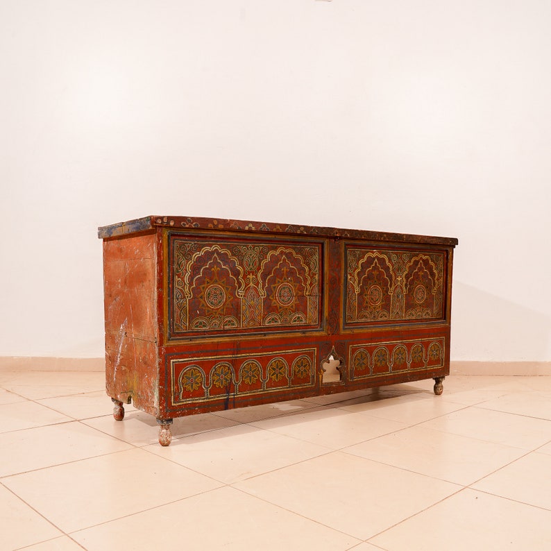 May include: A vintage wooden chest with intricate painted designs in shades of red, gold, and blue. The chest has two doors and a carved wooden base.