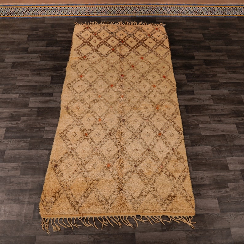 Pu&ograve; includere: Un tappeto marocchino beige con un motivo a rombi e accenti marroni. Il tappeto ha un bordo a frangia ed &egrave; fatto di lana.