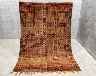 Puede incluir: Una alfombra marroquí vintage con un patrón geométrico en tonos rojos, marrones y dorados. La alfombra tiene un borde de flecos y está hecha de lana.