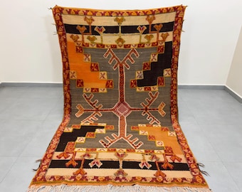 Puede incluir: Una alfombra vintage con un patrón geométrico en tonos naranja, rojo, marrón y blanco. La alfombra tiene un borde de flecos y está hecha de lana.