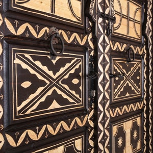 Op de afbeelding: Een donkerhouten kast met ingewikkelde houtsnijwerk en zwart metalen beslag. De kast heeft een geometrisch patroon met witte accenten en is versierd met pijlen en andere symbolen.