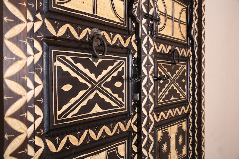 Op de afbeelding: Een donkerhouten kast met ingewikkelde houtsnijwerk en zwart metalen beslag. De kast heeft een geometrisch patroon met witte accenten en is versierd met pijlen en andere symbolen.