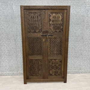 Op de afbeelding: Een donkerbruin houten kast met ingewikkelde gesneden ontwerpen op de deuren. De kast heeft twee deuren met een metalen grendel en een sleutelgat.