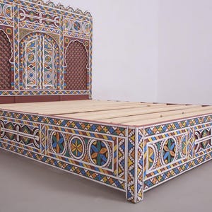 May include: A decorative bed frame with a headboard and footboard. The frame is painted with intricate geometric patterns in white, blue, yellow, and red. The headboard features arched designs and a textured brown panel.