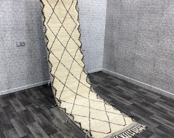 May include: A white and black Moroccan rug with a diamond pattern. The rug is hanging on a wall and has fringe on the bottom.