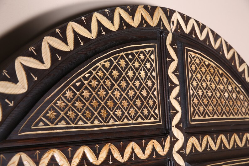 Op de afbeelding: Een donkerbruine houten deur met ingewikkelde houtsnijwerk in een geometrisch patroon. De houtsnijwerk is gemaakt van lichter hout en cre&euml;ert een contrastrijk ontwerp. De deur is gebogen aan de bovenkant en heeft een decoratieve rand.