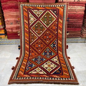 May include: A traditional Moroccan rug with a geometric pattern in shades of red, orange, blue, green, and white. The rug has a fringe border and is made of wool.
