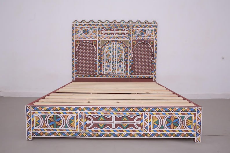 May include: A handcrafted bed frame with intricate, colourful geometric patterns. The headboard features a central arched design with a door motif, flanked by arched panels. The frame is painted in shades of red, blue, yellow, and white, creating a vibrant, decorative piece.