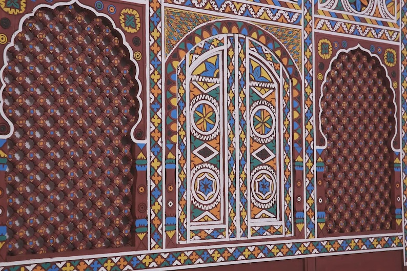 May include: A section of a wall with intricate, colourful geometric designs. The wall features a central arched doorway with detailed patterns in white, blue, yellow, and green. Flanking the doorway are arched panels with a repeating diamond pattern in a reddish-brown hue.