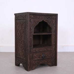 Peut inclure: Meuble en bois brun foncé avec des motifs floraux et géométriques sculptés. Il comprend un tiroir avec une poignée ronde, une étagère ouverte et un sommet en forme d'arche. Ce meuble est une unité de rangement décorative.