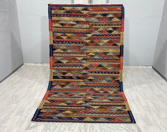 Könnte beinhalten: Ein bunter Teppich mit geometrischem Muster und Fransenbordüre. Der Teppich hat eine Mischung aus roten, blauen, gelben und braunen Farben.