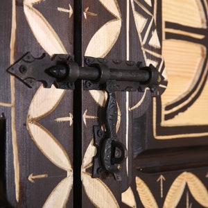 Op de afbeelding: Een close-up van een houten deur met ingewikkelde houtsnijwerk en een zwart metalen grendel. De deur is geschilderd in een donkerbruine kleur met witte accenten. De grendel is gemaakt van metaal en heeft een eenvoudig ontwerp.
