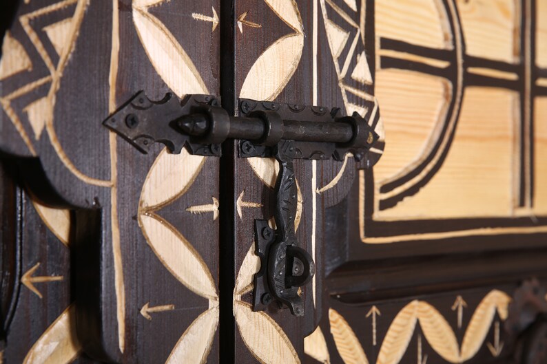 Op de afbeelding: Een close-up van een houten deur met ingewikkelde houtsnijwerk en een zwart metalen grendel. De deur is geschilderd in een donkerbruine kleur met witte accenten. De grendel is gemaakt van metaal en heeft een eenvoudig ontwerp.