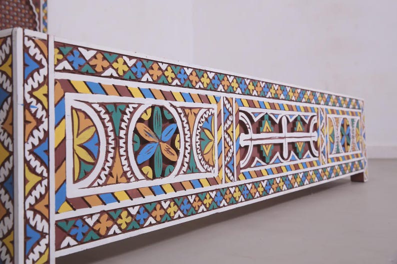 May include: A low-angle view of a white wooden bed frame featuring detailed, colourful geometric patterns. The design incorporates yellow, blue, orange, and brown hues, creating a vibrant aesthetic. The bed frame is a decorative piece.