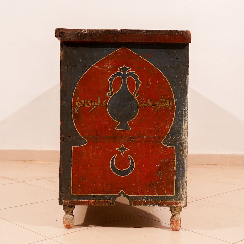 May include: A vintage wooden chest with a red, blue and gold painted design. The chest features an arched top with a crescent moon and star, and Arabic script.