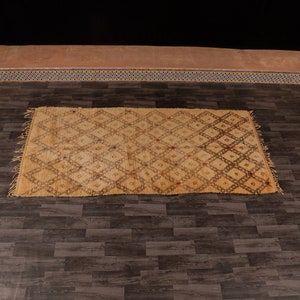 Pu&ograve; includere: Un tappeto marocchino beige con un motivo a rombi intrecciato in marrone. Il tappeto ha frange su tutti i lati.