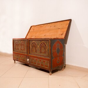 May include: A wooden chest with a hinged lid, painted in red, gold, and blue with intricate geometric patterns and Arabic script. The chest is decorated with a stylised vase and crescent moon.