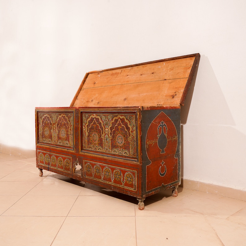 May include: A wooden chest with a hinged lid, painted in red, gold, and blue with intricate geometric patterns and Arabic script. The chest is decorated with a stylised vase and crescent moon.