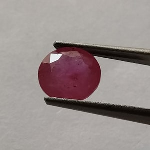 Könnte beinhalten: Ein ovaler Edelstein in tiefroter Farbe, der mit einer Pinzette gehalten wird.