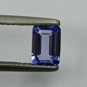 May include: A purple gemstone with a rectangular emerald cut. The gemstone is held by a pair of tweezers.