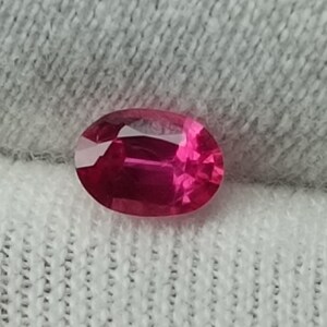 Puede incluir: Una vibrante gema de rub&iacute; de corte ovalado. La gema es de un rojo intenso y saturado con reflejos internos, sobre un fondo de tela blanca texturizada. La superficie pulida del rub&iacute; captura la luz, mostrando su brillo.