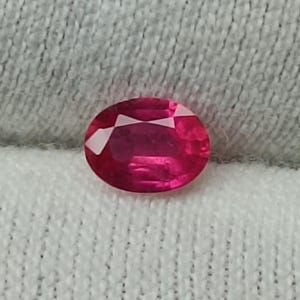 Könnte beinhalten: Ein oval geschliffener Rubin-Edelstein in tiefroter Farbe. Die Facetten des Steins reflektieren das Licht. Der Rubin liegt auf einem weißen, strukturierten Hintergrund, der seine Farbe hervorhebt. Geeignet für Schmuck.