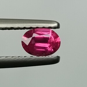 Puede incluir: Una vibrante gema rosa, de corte ovalado, es sostenida por unas pinzas plateadas sobre un fondo neutro. Las facetas de la gema reflejan la luz, mostrando su claridad y color. Esta imagen resalta el brillo de la gema.