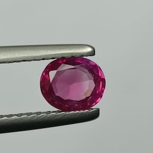 Peut inclure: Une gemme rose foncé, de forme ovale et taillée à facettes, est maintenue par une pince argentée. La pierre précieuse présente une coupe brillante. Le fond est gris neutre.