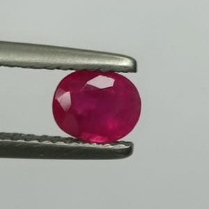 Peut inclure: Une pierre précieuse de rubis ovale et facettée, tenue par une pince. Le rubis est d'un rouge profond et vibrant, avec des facettes visibles qui captent la lumière. La pince est de couleur argentée.