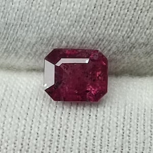 Könnte beinhalten: Ein facettierter, rechteckig geschliffener Rubin-Edelstein. Der Edelstein hat eine tiefe, satte rote Farbe mit einer leichten inneren Textur. Der Schliff weist scharfe, klare Linien und eine polierte Oberfläche auf, die das Licht reflektiert. Der Hintergrund ist weiß.