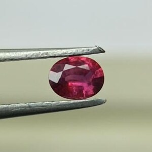 Könnte beinhalten: Nahaufnahme eines leuchtend roten, ovalen Edelsteins, der mit einer Pinzette gehalten wird. Der Edelstein ist ein Rubin, ein Edelstein, der für seine tiefrote Farbe bekannt ist.