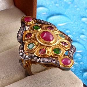 Könnte beinhalten: Ein goldener Ring mit einem großen Rubin in der Mitte, umgeben von kleineren Rubinen, Smaragden und Diamanten. Der Ring ist auf einem silbernen Band gefasst.