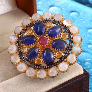 Könnte beinhalten: Ein goldener Ring mit einem runden Design, das eine Ansammlung von blauen und roten Edelsteinen zeigt, die von weißen Mondsteinen umgeben sind.