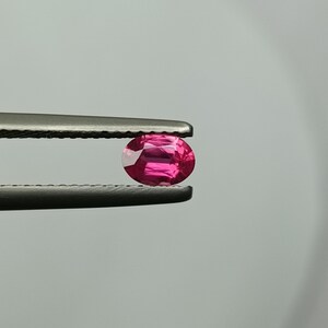 Puede incluir: Una vibrante gema roja rub&iacute; de corte ovalado, sujeta con unas pinzas plateadas. Las facetas de la gema reflejan la luz, mostrando su brillo. El fondo es gris neutro, resaltando el color y la claridad de la gema. Ideal para joyer&iacute;as.