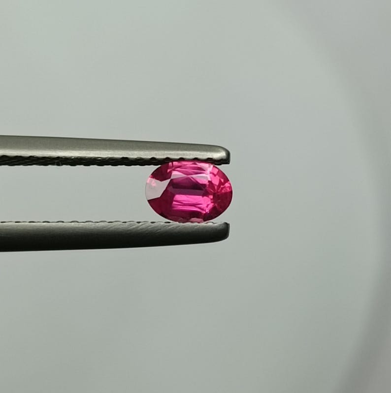 Puede incluir: Una vibrante gema roja rub&iacute; de corte ovalado, sujeta con unas pinzas plateadas. Las facetas de la gema reflejan la luz, mostrando su brillo. El fondo es gris neutro, resaltando el color y la claridad de la gema. Ideal para joyer&iacute;as.