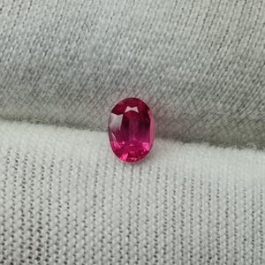 Puede incluir: Un rub&iacute; de talla ovalada, de color rojo-rosa intenso. La gema muestra reflejos internos y est&aacute; colocada sobre una tela blanca texturizada. La forma del rub&iacute; es alargada.