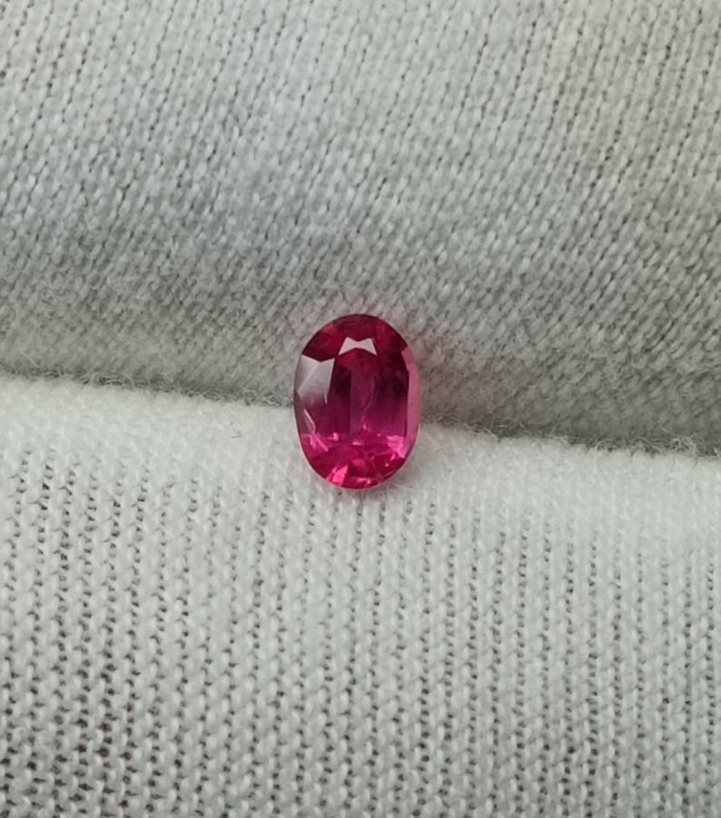 Puede incluir: Un rub&iacute; de talla ovalada, de color rojo-rosa intenso. La gema muestra reflejos internos y est&aacute; colocada sobre una tela blanca texturizada. La forma del rub&iacute; es alargada.