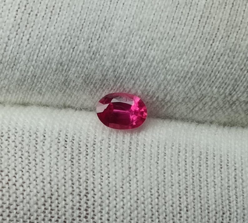 Puede incluir: Una vibrante gema de rub&iacute; de corte ovalado. La gema es de un rojo intenso y saturado con reflejos internos, sobre un fondo de tela blanca texturizada. La superficie pulida del rub&iacute; captura la luz, mostrando su brillo.
