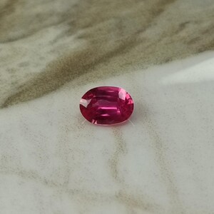 Puede incluir: Una gema de rub&iacute; de corte ovalado. La gema es de un color rojo-rosa intenso y saturado, con facetas que capturan la luz. El rub&iacute; se presenta sobre un fondo neutro, resaltando su brillo y claridad. Una hermosa gema para joyer&iacute;a.