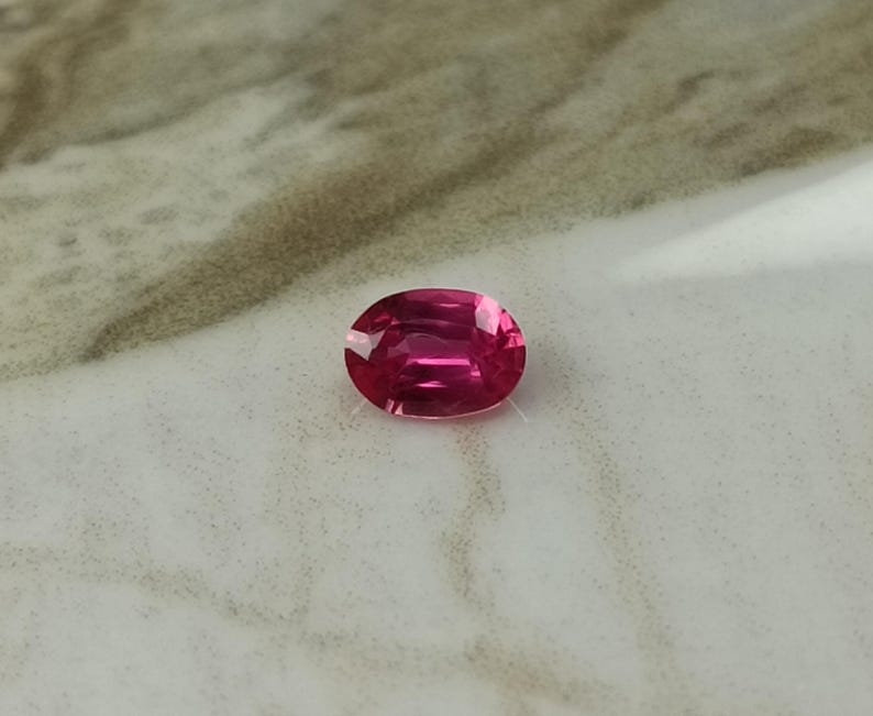 Puede incluir: Una gema de rub&iacute; de corte ovalado. La gema es de un color rojo-rosa intenso y saturado, con facetas que capturan la luz. El rub&iacute; se presenta sobre un fondo neutro, resaltando su brillo y claridad. Una hermosa gema para joyer&iacute;a.