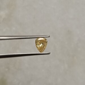 May include: A pear-shaped, yellow diamond gemstone held by tweezers.