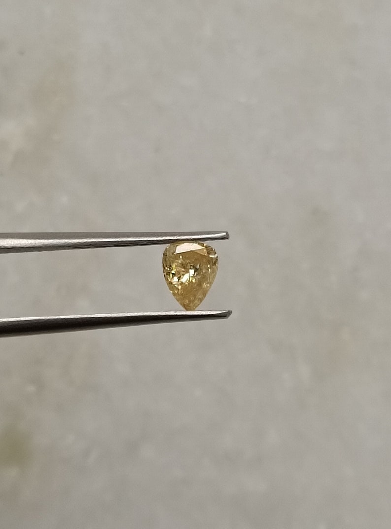 May include: A pear-shaped, yellow diamond gemstone held by tweezers.