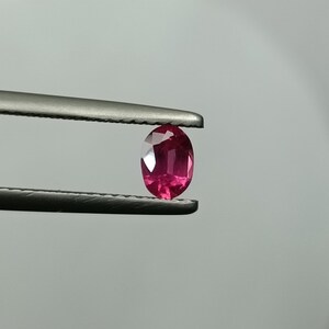 Puede incluir: Un rub&iacute; de forma ovalada, sujeto con unas pinzas. La gema presenta un color rosa intenso y facetas que capturan la luz. El rub&iacute; se muestra sobre un fondo neutro, resaltando su claridad y brillo. Las pinzas son met&aacute;licas.