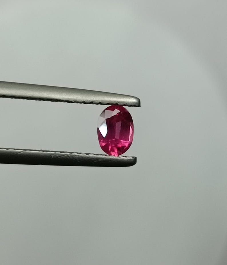Puede incluir: Un rub&iacute; de forma ovalada, sujeto con unas pinzas. La gema presenta un color rosa intenso y facetas que capturan la luz. El rub&iacute; se muestra sobre un fondo neutro, resaltando su claridad y brillo. Las pinzas son met&aacute;licas.